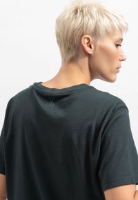 T-shirt in cotone verde scuro con collo a giro e maniche corte. Presenta una piccola grafica di forbici all'altezza del collo dietro. Texture liscia.