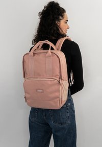 Zaino in tessuto rosa con manici superiori duali, spallacci imbottiti, tasca frontale con zip e dettagli testurizzati. Design pulito e rettangolare.