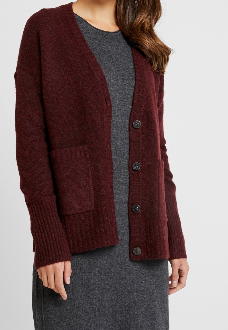 Burgundowy sweter w splot z dekoltem w serek, ściśle dopasowane mankiety, dwie przednie kieszenie oraz zapięcie na czarne guziki, noszony na szarej sukience.