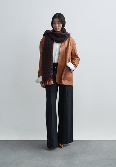 Brun läderjacka, vit skjorta, marinblå vida byxor och en mönstrad mörkröd scarf med fransar. Leopardskinnsklackar synliga.