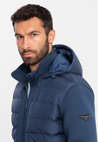 Marinblå puffjacka med huva, med quiltad textur, ribbat krage och metallisk logotypdetalj på axeln.