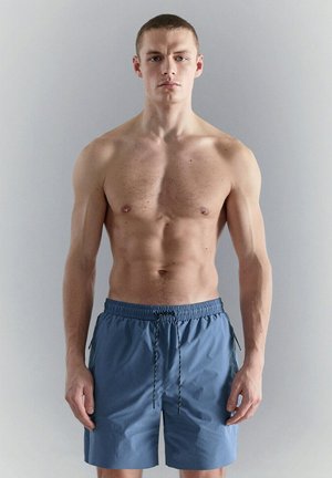 Giovane uomo con capelli corti in piedi, senza maglietta, che indossa pantaloncini da bagno blu con coulisse su uno sfondo grigio chiaro uniforme.