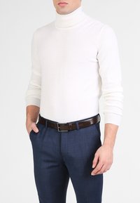 Pull col roulé blanc en matière douce, associé à un pantalon texturé bleu foncé et une ceinture en cuir marron avec une boucle en argent.