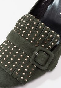 Mocassin en daim vert foncé avec ornements cloutés dorés, empeigne plissée et bride à boucle assortie en daim.