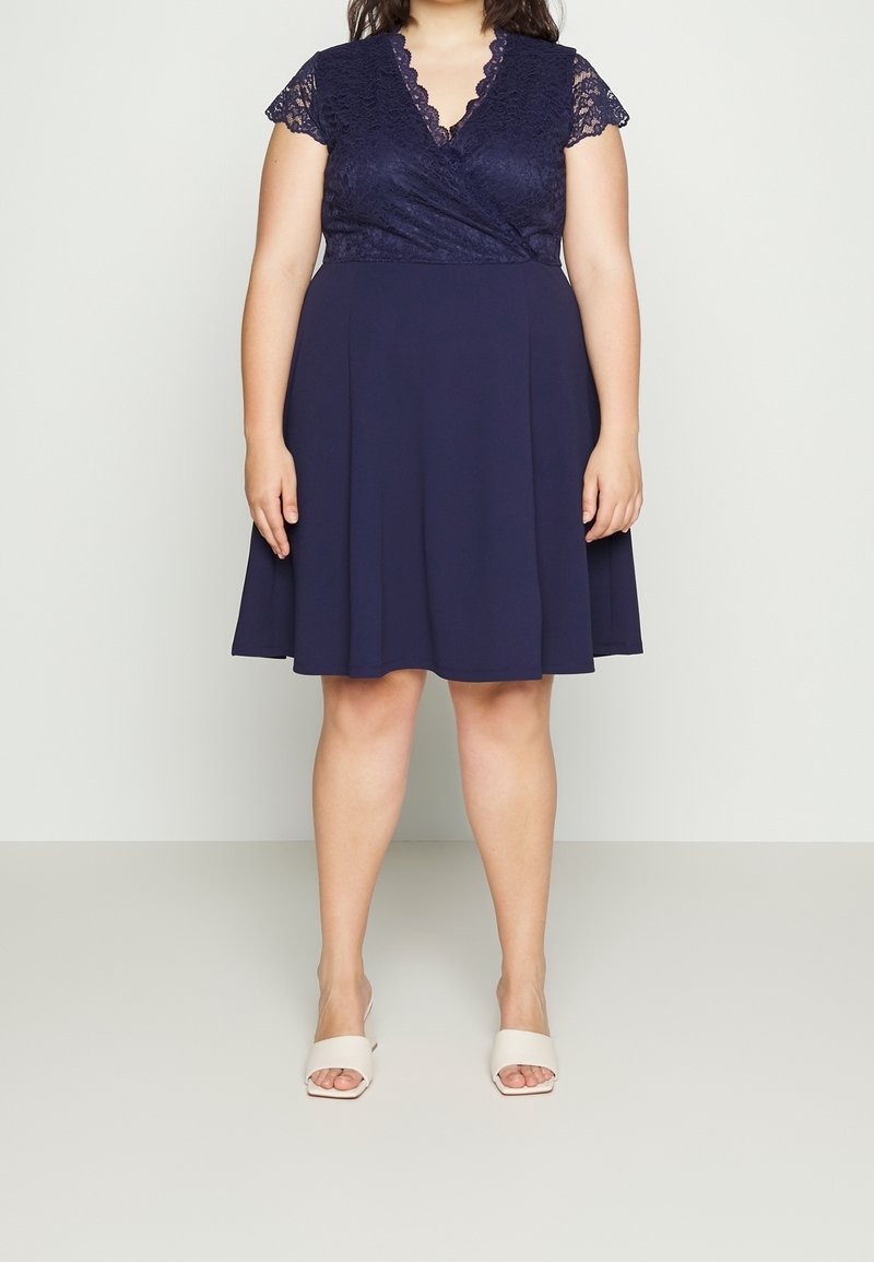 Femme portant une robe bleu marine longueur genou avec manches courtes en dentelle et col en V, associée à des sandales blanches à talons ouverts.