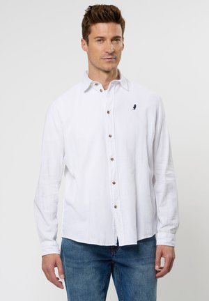 Camisa de lino blanca de manga larga con botones en la parte delantera, botones marrones y un pequeño logo azul en el pecho. Combinada con jeans azules.