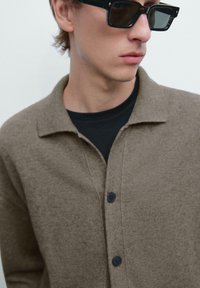 Brun stickad cardigan med krage och blå knappar, med en strukturerad yta, lagd över en svart T-shirt.
