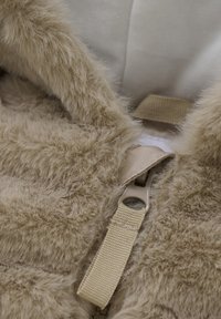 Beige nepbontproduct met een zachte textuur, gedetailleerde stiksels, een beige band en een plastic rits met een brede trekker.