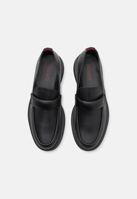 Bottines noires à enfiler avec une tige en cuir lisse, un bout rond et une sangle surélevée sur le cou-de-pied. Présente une semelle en caoutchouc texturée.