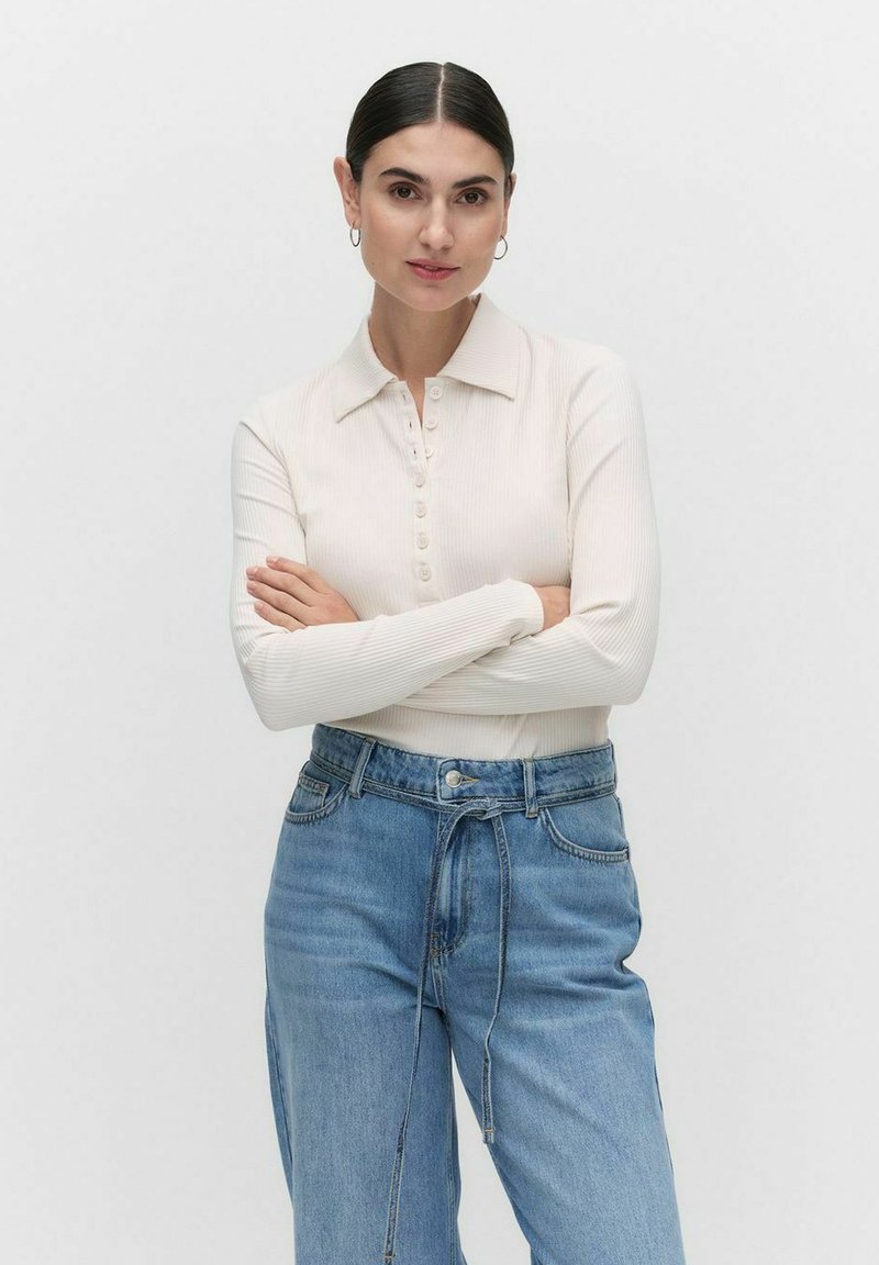 Pitkähihaisessa kerma värisessä ribbipaidassa on kaulus ja nappilista, ja se on yhdistetty löysiin sinisiin farkkuihin, joissa on nyörivarustus vyötäröllä.