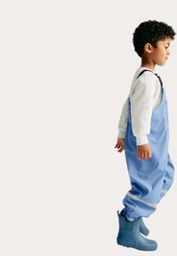 Bambino che indossa una felpa grigia chiara, salopette blu impermeabili con strisce riflettenti e stivali da pioggia blu abbinati, in piedi di lato.