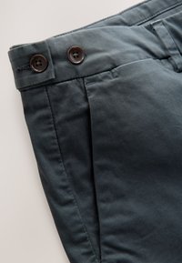 Tamsiai žalių kelnių priekinė dalis plokščia, juosmens diržo vietoje du rudi mygtukai ir matomos siuvimo linijos. Pagamintos iš lygaus audinio.