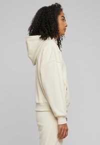 Hoodie en polaire crème avec une capuche à cordon de serrage, une grande poche avant, des poignets et un ourlet côtelés. Présente une coupe décontractée et une texture douce.