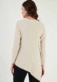 Maglietta beige a maniche lunghe con un motivo texturizzato, caratterizzata da un orlo curvo e fessure laterali. Indossata con pantaloni neri, presenta una silhouette rilassata.