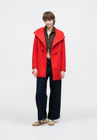 Red wool coat with wide lapels, double-breasted buttons, and side pockets; paired with dark wide-leg jeans and patterned sneakers.