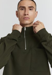 Mørkegrøn zip-neck sweatshirt med lange ærmer, der har en brystlomme og ribbede manchetter. Blødt stof med et minimalistisk design.