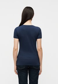 Γυναίκα με μακριά σκούρα μαλλιά, ντυμένη με στενό μπλε σκούρο κοντομάνικο πουκάμισο και σκούρο τζιν, που στέκεται με την πλάτη στην κάμερα.