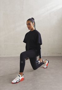 Haut de sport noir et leggings, chaussures de course à motif avec des accents orange. La personne effectue une fente en tenant un haltère.