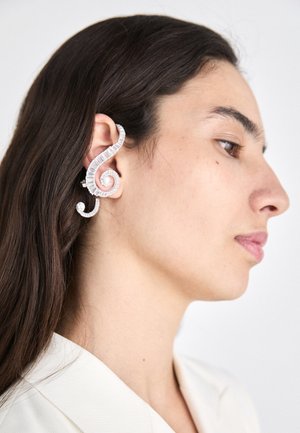 Orecchino a clip in argento con un design a spirale, superficie texturizzata e forma curva, indossato all'orecchio sinistro contro capelli scuri e lisci.