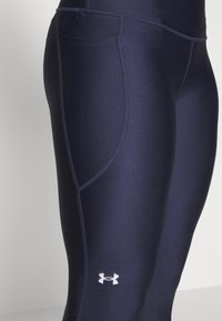 Granatowe legginsy sportowe z gładkiego, elastycznego materiału, z boczną kieszenią, tylnym przeszyciem i odblaskowym logo na dolnej części nogi.