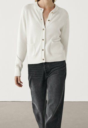 Kvinde iført en hvid cardigan med knapper og to forlommer samt sorte højtaljede jeans mod en ensfarvet baggrund.