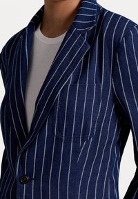 Navy blue striped blazer featuring vertical white stripes, peak lapels, a single chest pocket, and a button closure. Soft textured fabric.