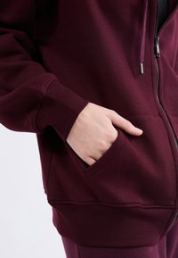 Felpa con zip color bordeaux, dotata di tasca frontale, polsini a coste e una consistenza morbida. Presenta una zip argentata e dettagli di cucitura a contrasto.