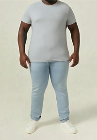 Homme portant un t-shirt gris clair ajusté, un jean bleu clair et des baskets blanches, debout devant un fond neutre.