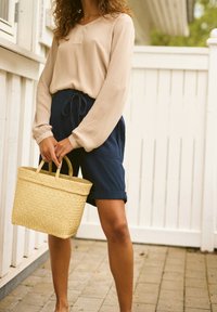 Haut beige à manches longues avec un petit plastron devant, associé à un short bleu marine et un sac panier en paille tressée, doté de deux anses.