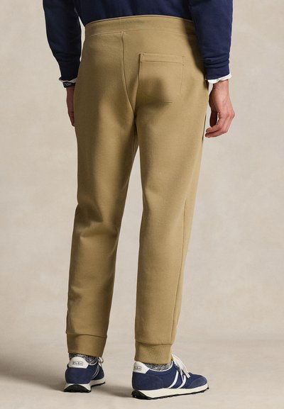 Man met beige joggingbroek en blauwe sneakers, staat met zijn rug naar de camera voor een effen achtergrond.