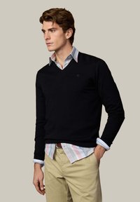 Mand med brunt hår iført sort V-hals trøje over pastelstribet skjorte med knapper og beige bukser, står med den ene hånd i lommen.