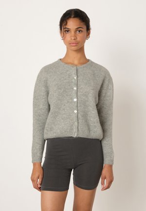 Naine, kes kannab helehallist nööpidega kardigani ja musti jalgrattapükse, seisab valge tausta ees.