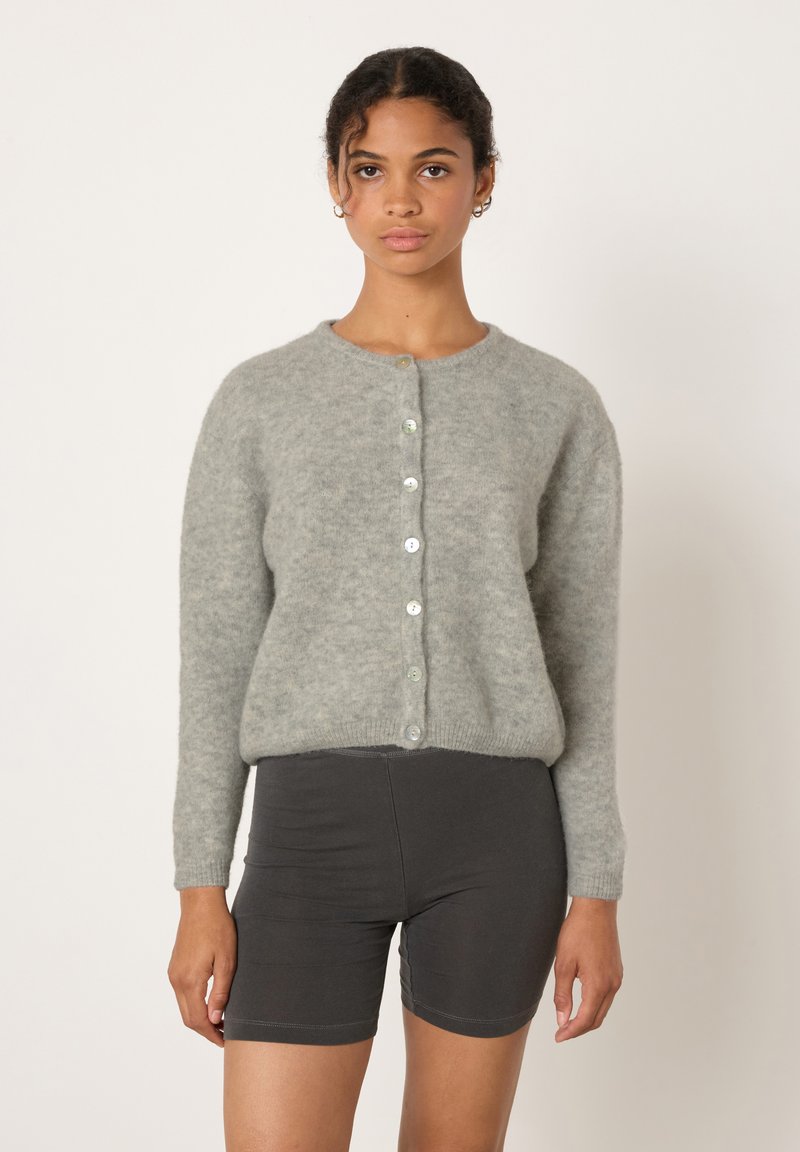 Moteris dėvi šviesiai pilką, užsegamą megztinį ir juodus dviračių šortus, stovinti priešais baltą foną.