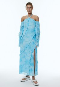 Vloeiende blauwe off-shoulder jurk met geruffelde randen en een zijsplit. Gemaakt van lichtgewicht, doorschijnende stof met een draaiend patroon.