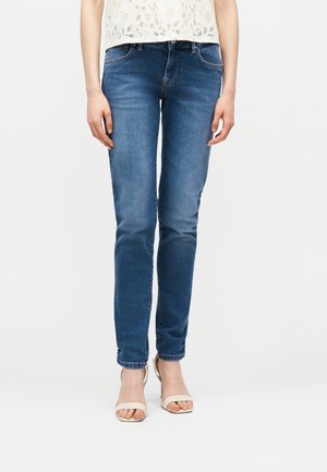 Donna che indossa jeans aderenti blu, top bianco in pizzo e tacchi beige aperti in punta, in piedi contro uno sfondo bianco semplice.