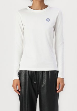 Personne portant une chemise blanche à manches longues avec un petit logo rond bleu souriant sur la poitrine et un pantalon en cuir noir ample.