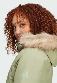 Světlé zelené sako s kožešinovým lemem na kapuci, s hladkým texturou a logem Adidas. Design má přiléhavý střih a zvýšený límec.