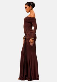 Lang brun kjole med off-shoulder-udskæring, lavet af et glat stof, med plisserede detaljer og en draperet design langs siden.