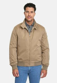 Chaqueta bomber marrón claro con cremallera frontal, puños y cuello acanalados, hecha de tela suave. Lleva puesta sobre una camisa de cuadros.