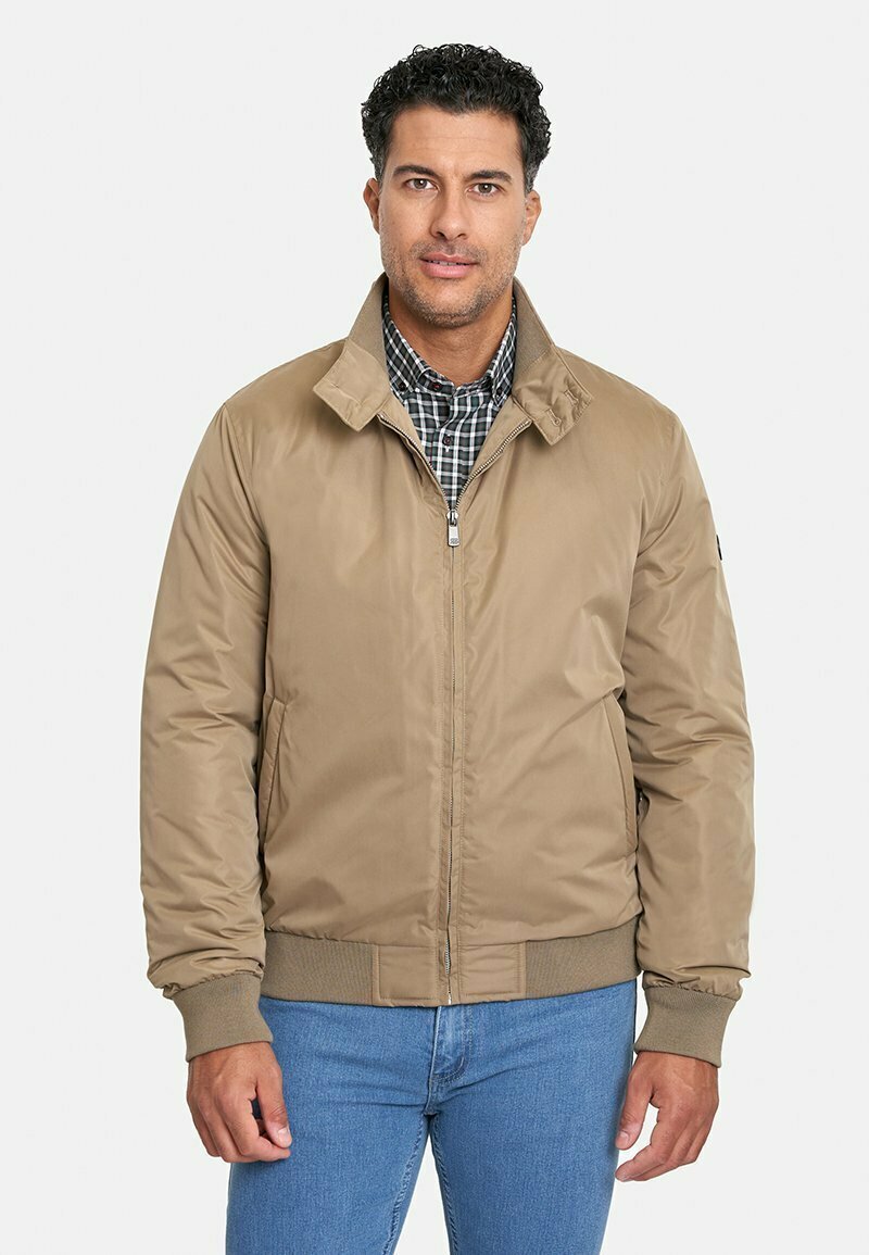 Chaqueta bomber marrón claro con cremallera frontal, puños y cuello acanalados, hecha de tela suave. Lleva puesta sobre una camisa de cuadros.