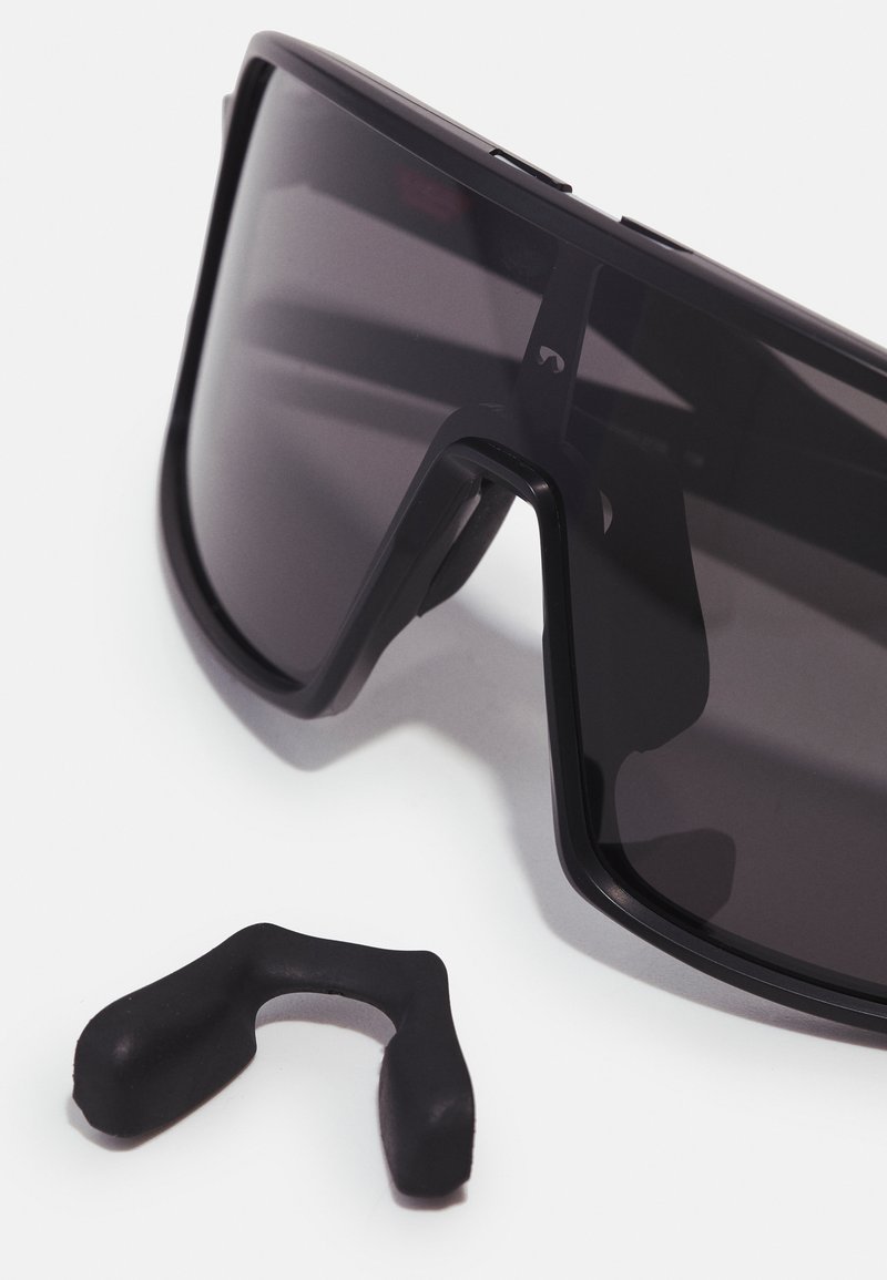 Lunettes de soleil noires au design enveloppant et aux verres teintés foncés. Comprend une pièce nasale en caoutchouc noir séparée. Texture lisse.