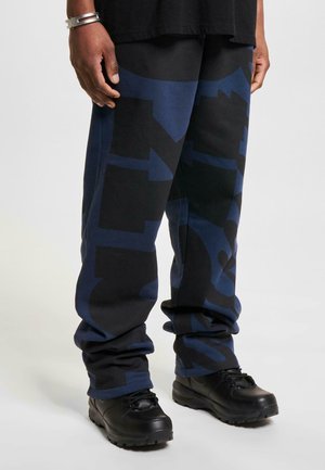 Blue and black fleece jogger pants with abstract patterns. Features cuffed hems, a relaxed fit, and paired with black, rugged shoes.