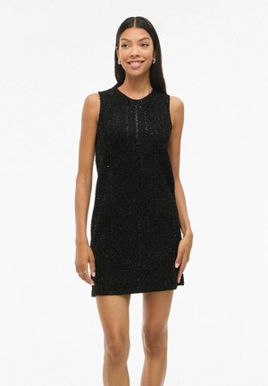 Mladá žena s dlouhými tmavými vlasy, oblečená v černých texturovaných minišatech bez rukávů, usmívající se, stojící před jednoduchým bílým pozadím.