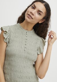 Haut texturé vert clair avec des manches courtes à volants et un col rond. Présente une patte de boutonnage avec de petits boutons assortis.