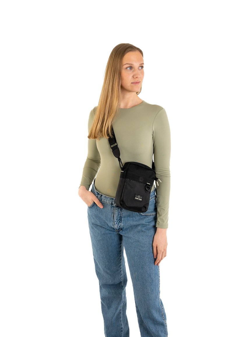 Borsa a tracolla in nylon nero con tracolla regolabile, tasca frontale e dettagli in metallo, indossata da un modello con un top attillato a maniche lunghe e jeans.