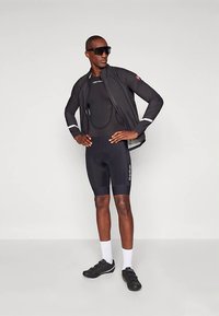 Juoda dviračių apranga su prigludusiu pagrindiniu sluoksniu ir šortais. Lengva juoda striukė su atšvaitiniais akcentais ir akiniai nuo saulės. Juodi dviračių batai.