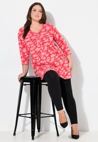 Czerwona kwiecista tunika z rękawami o długości trzech ćwierć, z detalami w postaci guzików, noszona na czarnych legginsach i czarnych szpilkach; siedząc na czarnym stołku.