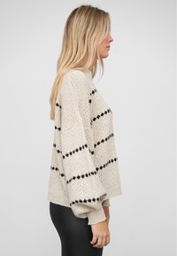 Sweter dzianinowy w beżowym kolorze z czarnymi poziomymi paskami, luźny krój, opadające ramiona i ściągacze. Teksturowana tkanina, styl casual.