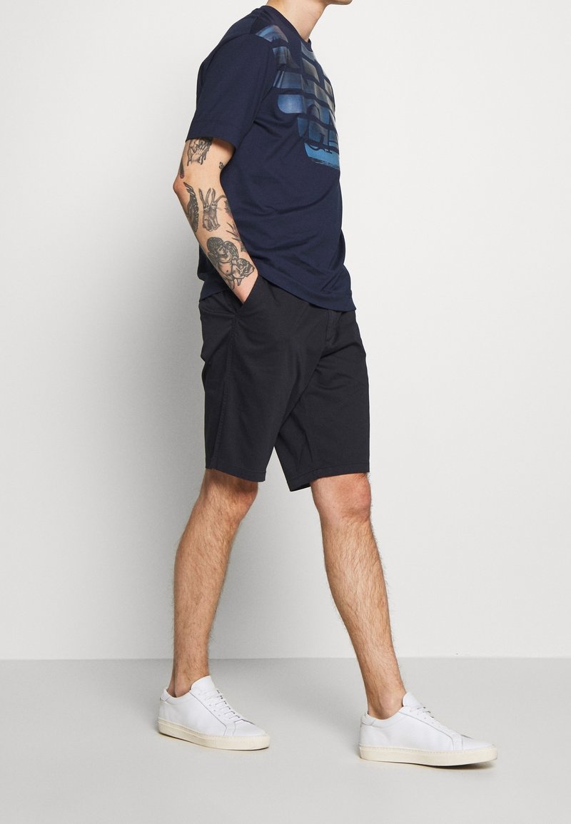 Marinblå t-shirt med grafisk design, matchat med svarta shorts. Modellen bär vita sneakers. Tyget ser mjukt ut; avslappnad passform och stil.