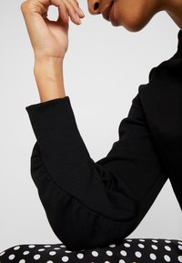 Musta pitkähihaisen puseron olkapäällä oleva täytetty muotoiluelementti ja istuva hihansuu. Taustalla on kuvioitu kangas, jossa on valkoisia pilkkuja.